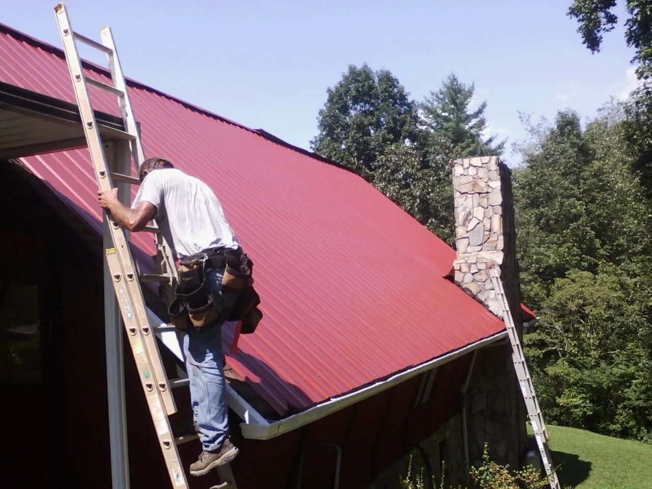 Roofer on a ladder beside a red metal roof and stone chimney — Soffit & Fascia Repair by Roofing Brothers in Rollinsville